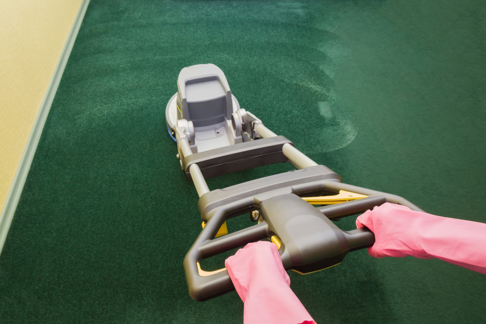 A person wearing pink gloves operates a carpet cleaning machine over a green carpet, visibly removing a section of dirt.